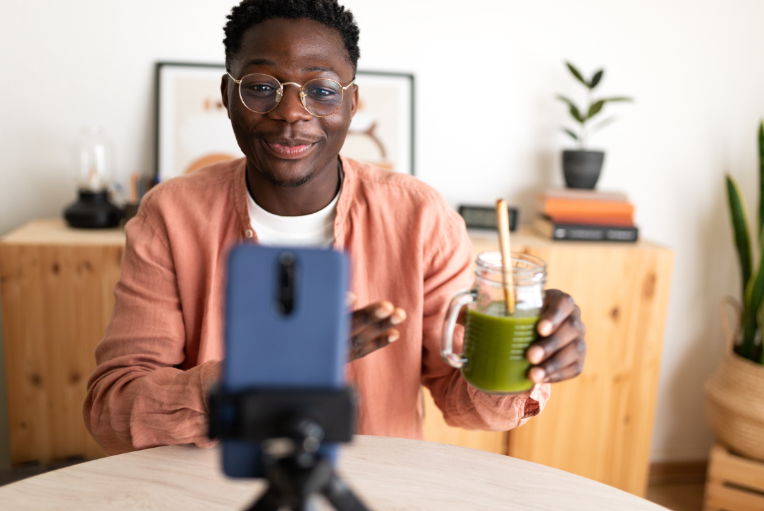 A man holds a smoothie and looks into a phone camera, which is set up on a tripod.