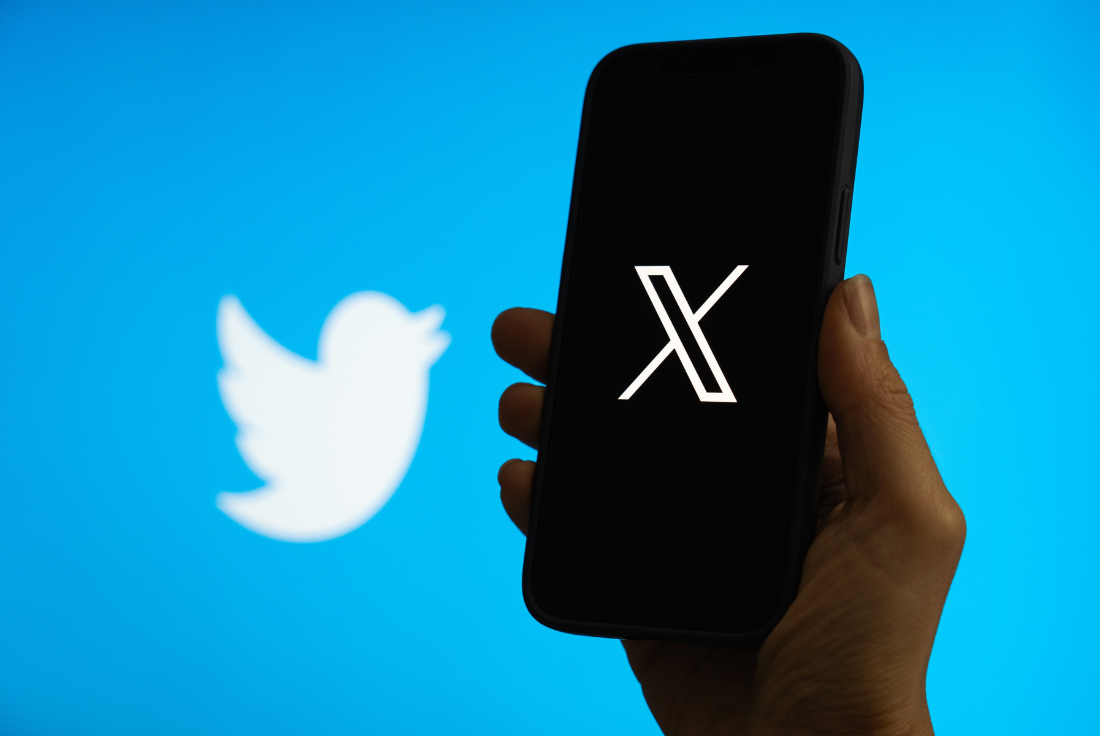 A person holds a phone with X's logo on it. The Twitter logo is on a screen behind it.