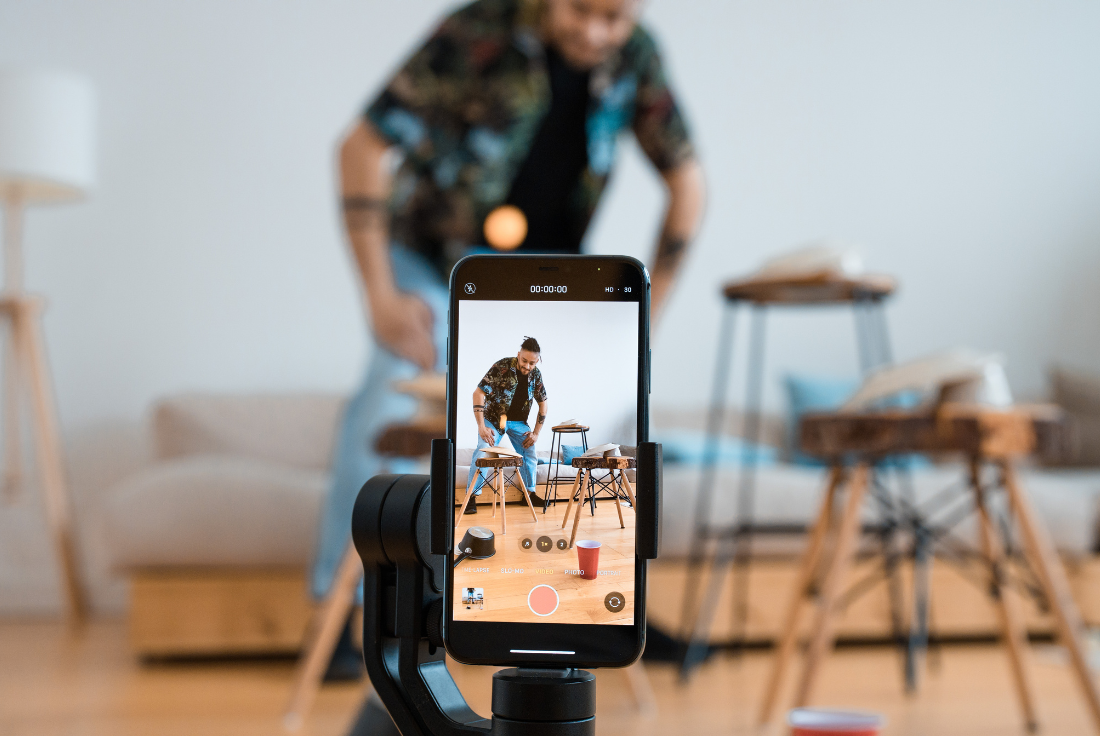 A phone on a tripod records a man trying to complete a trick shot with a ping pong ball.