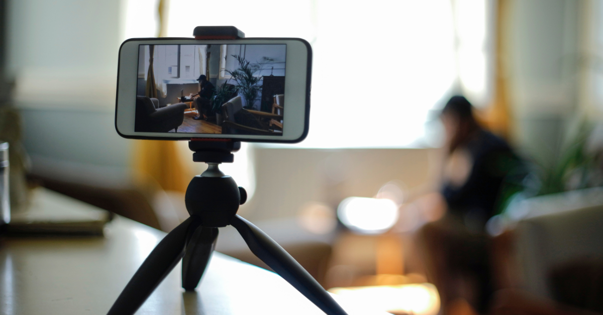 A phone, open to the camera app, sits on a tripod on a shelf. The camera is pointed at a person sitting in a chair, typing on a laptop. 